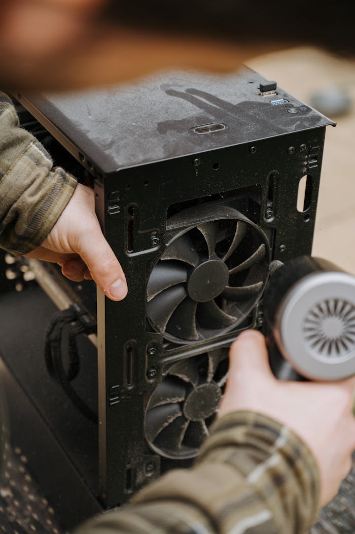 Hands cleaning dust from a computer fan using an electric air duster for maintenance.